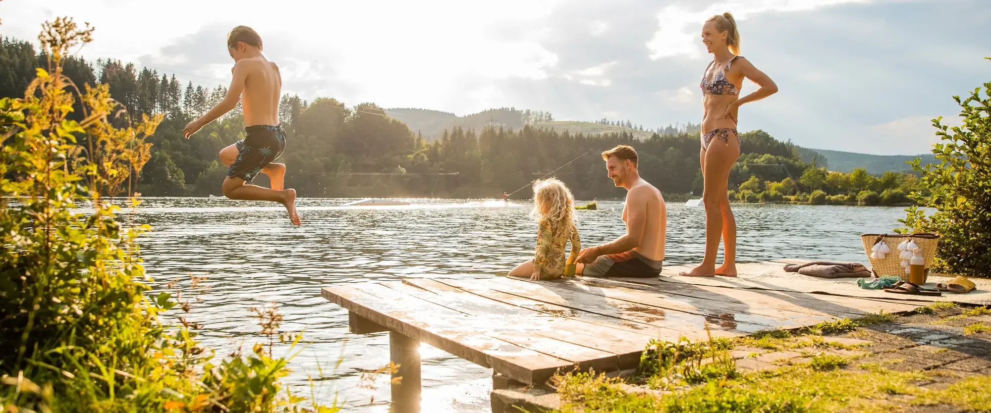 Eine Familie mit zwei Kindern, von denen eines ins Wasser springt, sitzt auf einem Steg.