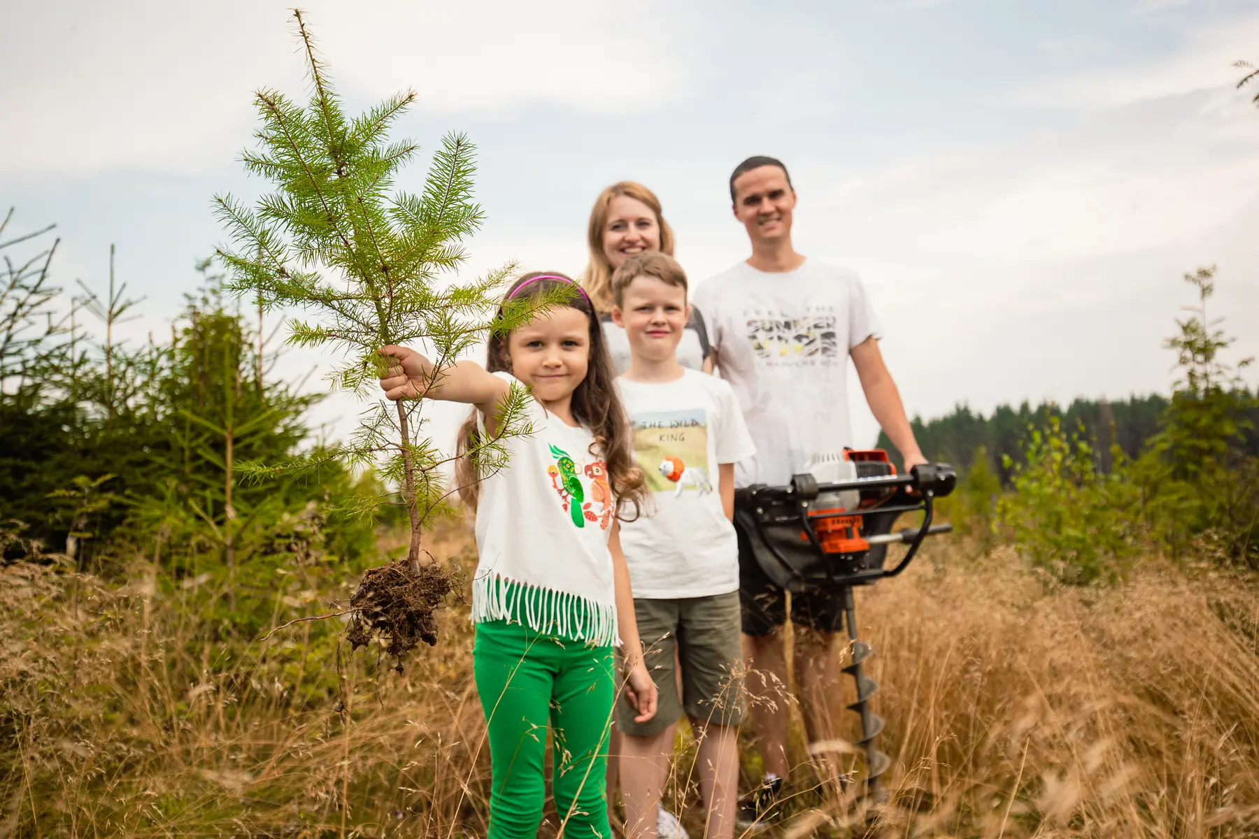 Eine Familie mit zwei Kindern will einen neuen Baum einpflanzen, den das kleine Mädchen noch in der Hand hält.