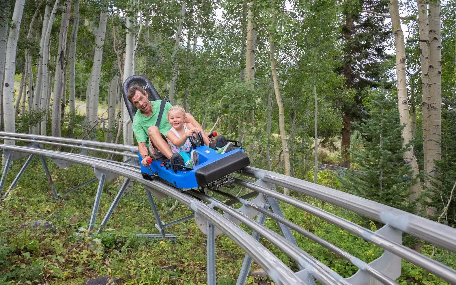 Ein Mann und ein Kind die auf einem Schlitten die Sommerrodelbahn herunterfahren und in die Kamera lächeln.