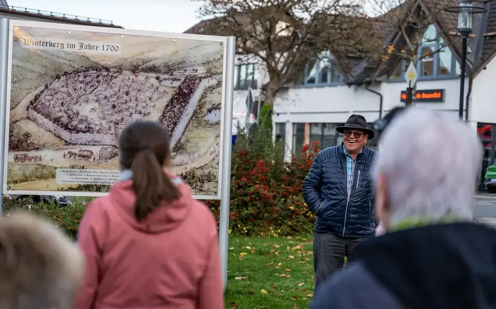 Mann erklärt vor einer Tafel mit einem abgebildeten Plan der Altstadt einer Gruppe etwas.