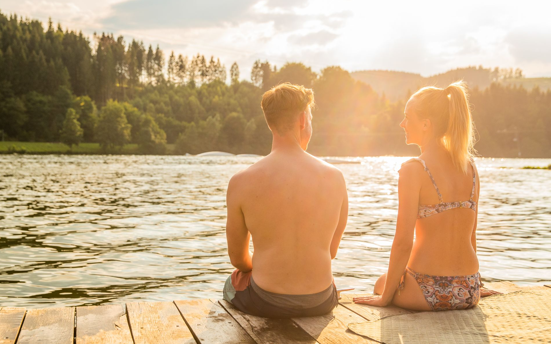 Ein Pärchen sitzt im Sonnenschein am Rande eines Stegs und lässt die Füße ins Wasser baumeln.