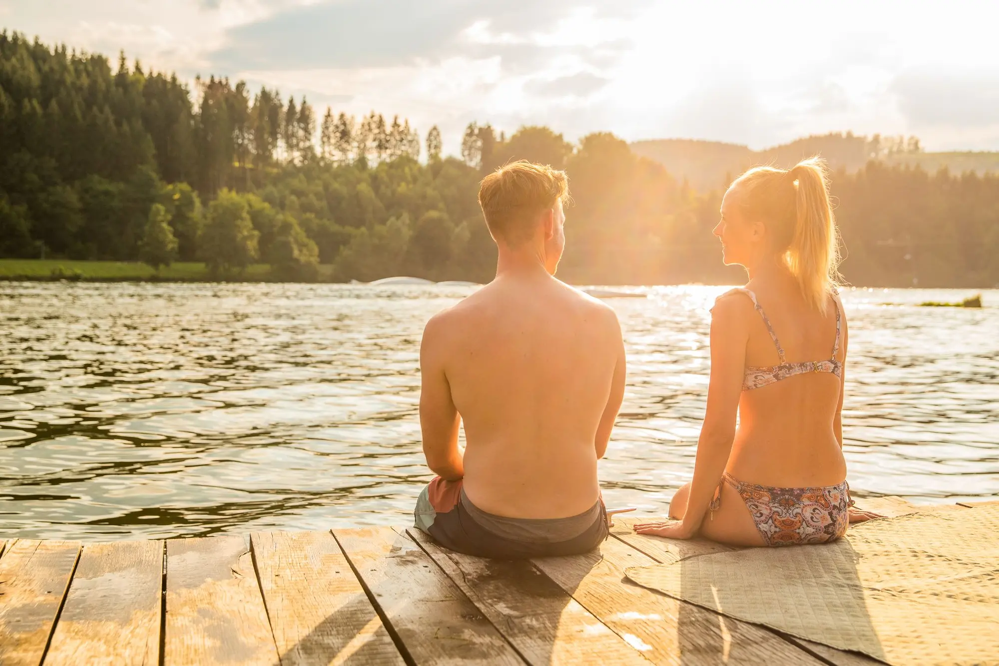 Ein Pärchen sitzt im Sonnenschein am Rande eines Stegs und lässt die Füße ins Wasser baumeln.