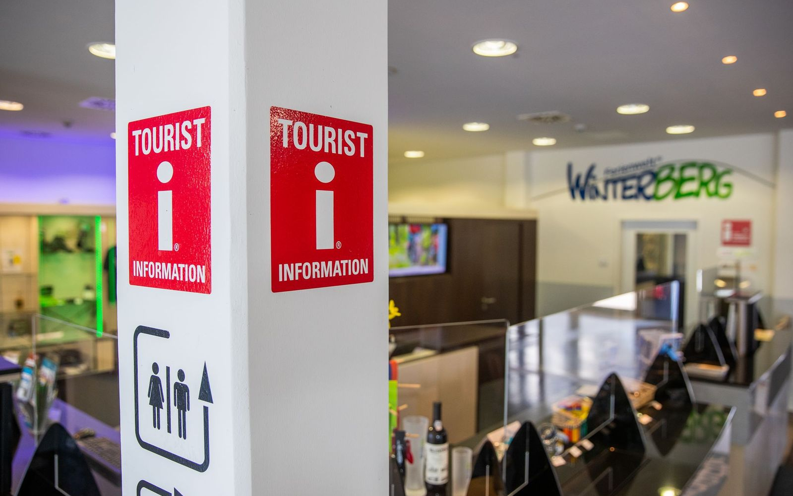 Das Logo für Tourist Informationen an einem Pfeiler mit dem Counter der Tourist Information im Hintergrund.