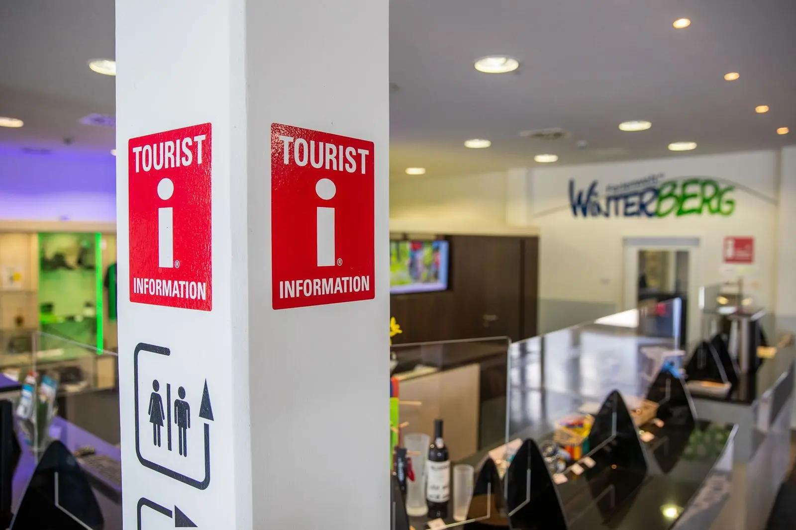 Das Logo für Tourist Informationen an einem Pfeiler mit dem Counter der Tourist Information im Hintergrund.