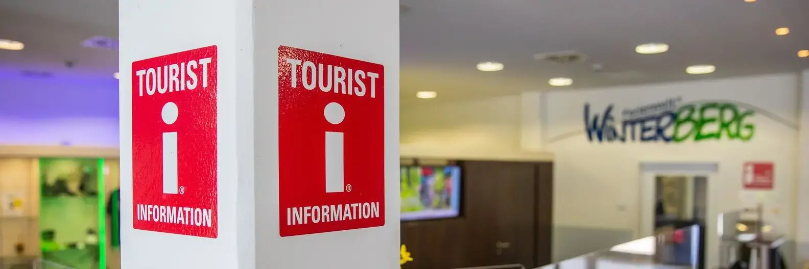 Das Logo für Tourist Informationen an einem Pfeiler mit dem Counter der Tourist Information im Hintergrund.