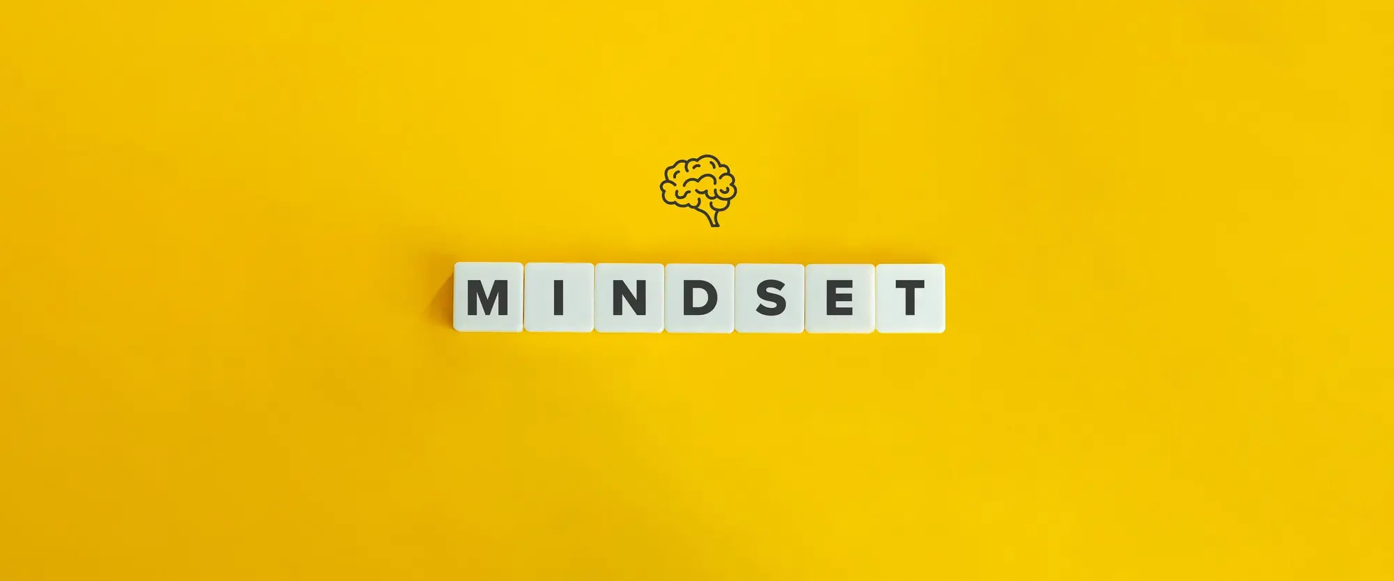 Das Wort Mindset aus Buchstabensteinen gelegt auf einem gelben Hintergrund. Darüber die Zeichnung eines Gehirns.