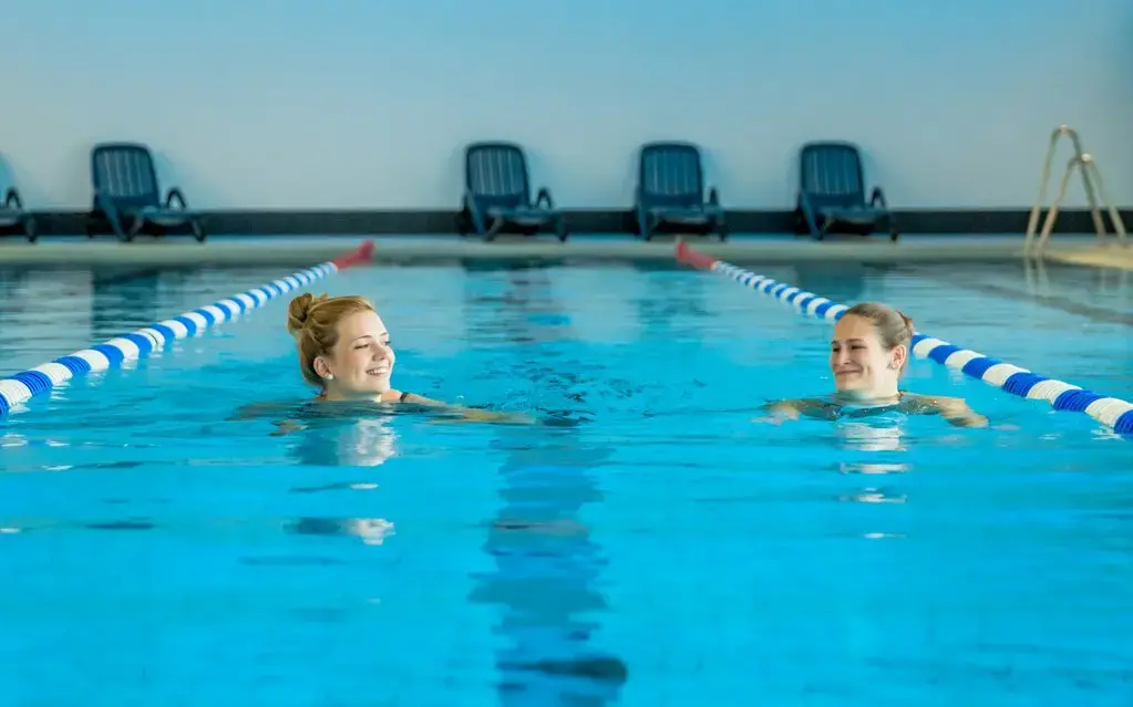 Zwei Frauen, die im Sportbecken nebeneinander her schwimmen und sich anlächeln.
