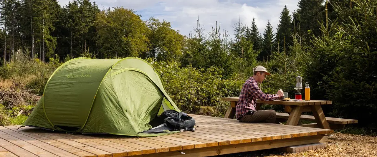 Ein Zelt auf einem hölzernen Podest aufgebaut. Daneben ein Mann auf einer Bank, der sein Essen genießt.