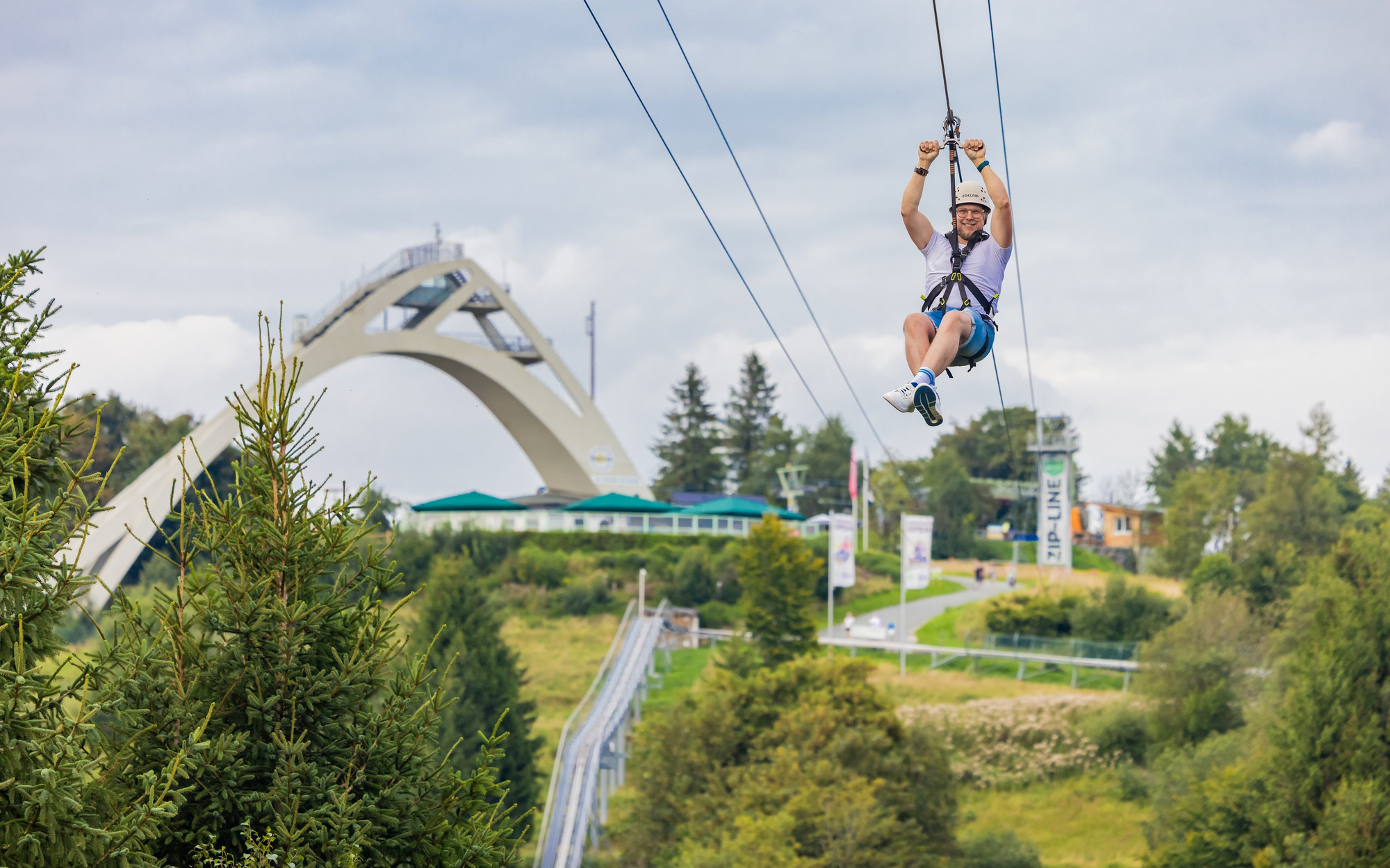 Ein Mann fliegt im Vordergrund der St. Georg Schanze die Zipline hinunter.