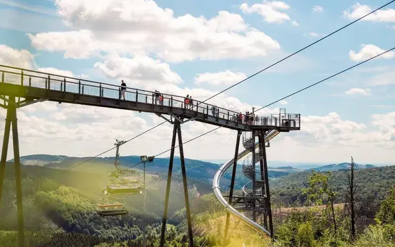 Mehrere Personen stehen am Ende der Panorama Erlebnisbrücke. Unter ihnen fährt ein Skilift nach oben und am Ende der Brücke sind eine Rutsche und eine Wendeltreppe zu sehen.