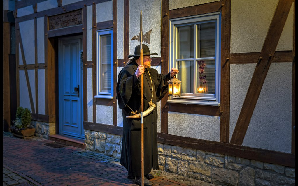 Ein Mann im Nachtwächteroutfit mit Hut und Mantel steht mit Laterne und Lanze in den Händen vor einem Fachwerkhaus und schaut Richtung Fenster.