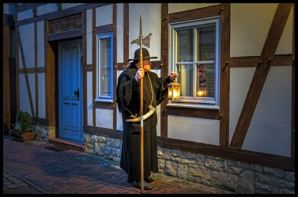 Ein Mann im Nachtwächteroutfit mit Hut und Mantel steht mit Laterne und Lanze in den Händen vor einem Fachwerkhaus und schaut Richtung Fenster.