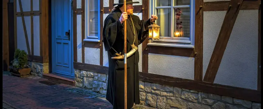 Ein Mann im Nachtwächteroutfit mit Hut und Mantel steht mit Laterne und Lanze in den Händen vor einem Fachwerkhaus und schaut Richtung Fenster.